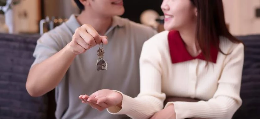 A smiling couple holding a house key with a miniature house keychain, marking the timeline for buying a house after reservation.